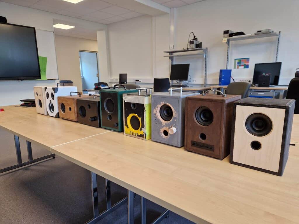 Elevproduserte boostere Åtte håndlagde trehøyttalere står på rekke og rad på et bord i et klasserom eller et verksted, med pulter, stoler og datamaskiner synlige i bakgrunnen.
