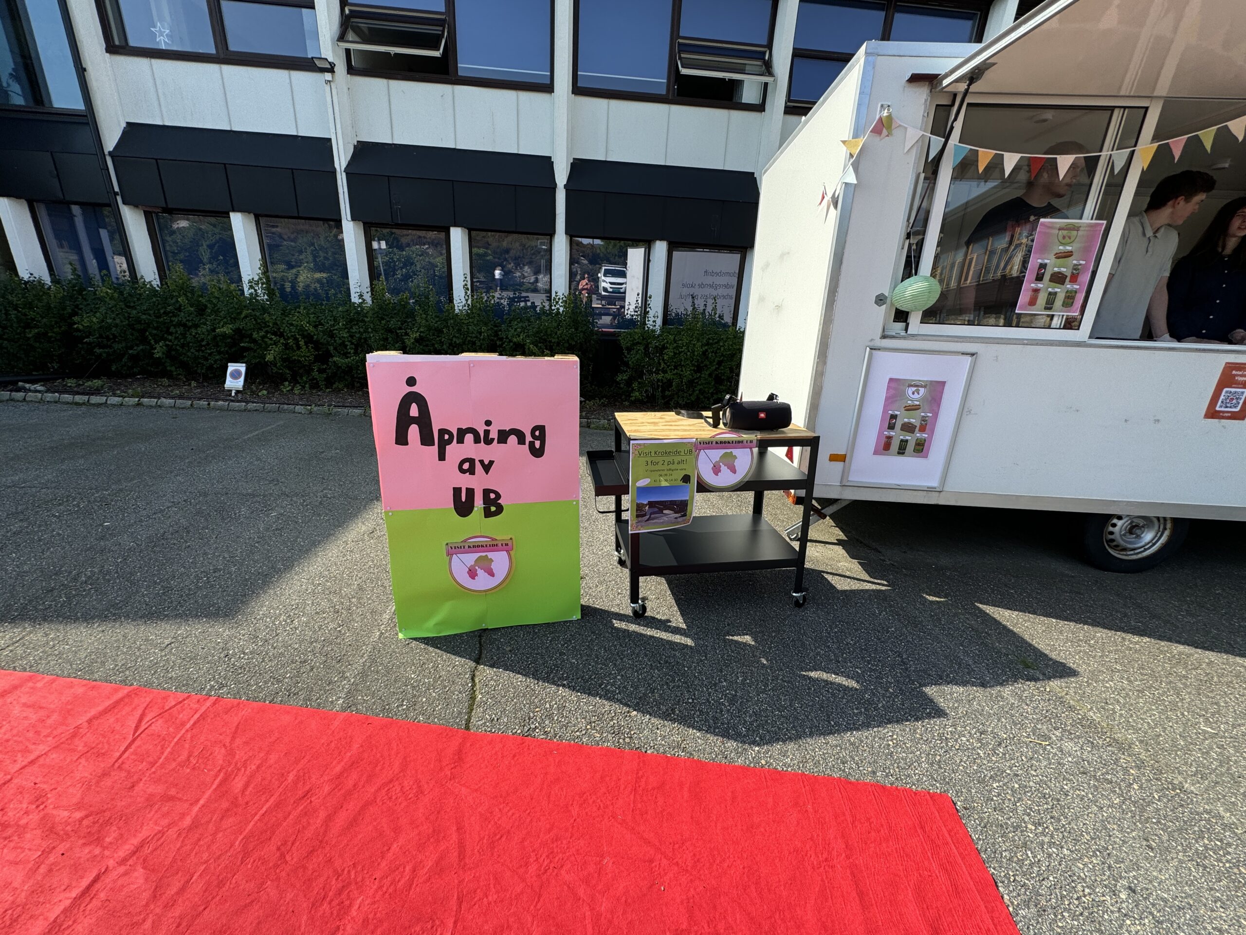 Et skilt med "Åpning av UB" står ved siden av en vogn og en hvit bod utenfor en bygning, med et rødt teppe på bakken.