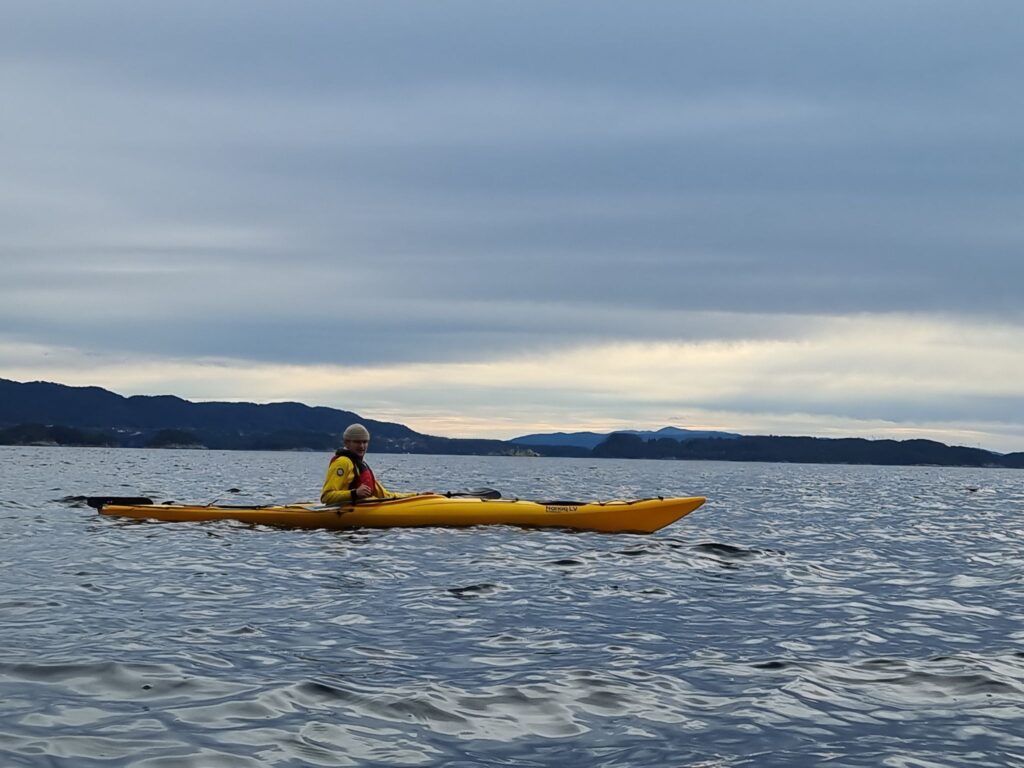 312907583_505106048330620_2413456327564104741_n En person i en gul kajakk padler på et stille vann under en overskyet himmel, med fjerne fjell synlige i bakgrunnen.