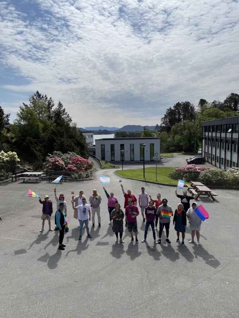 345125280_1574613533029876_5109452493863528704_n-scaled En gruppe mennesker står ute på et asfaltert område med ulike prideflagg i hendene, med bygninger, grøntområder og en delvis overskyet himmel i bakgrunnen.