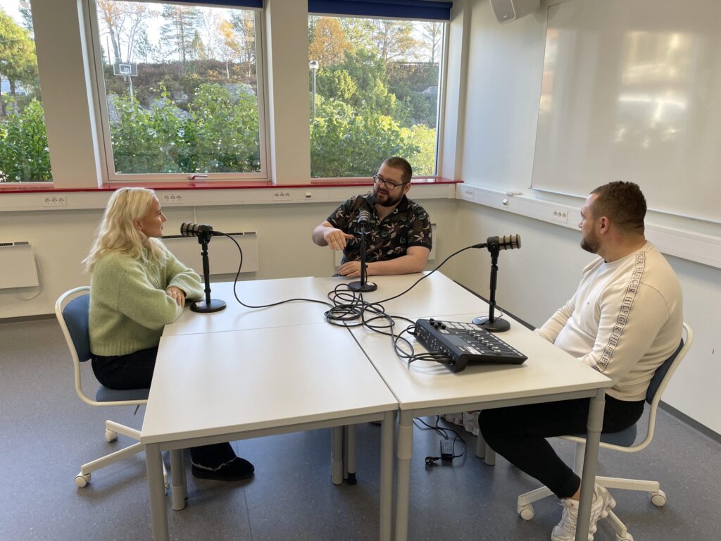 Tre personer sitter rundt et bord med mikrofoner og lydutstyr og spiller inn en podkast i et lyst rom med store vinduer.