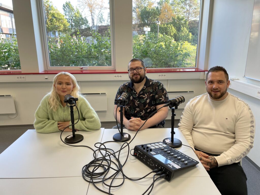 Tre personer sitter ved et bord med mikrofoner og lydutstyr og ser ut til å spille inn en podkast eller et intervju i et lyst rom med store vinduer og trær utenfor.