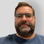 En mann med briller og skjegg iført blå genser poserer foran en vanlig, lysegrå bakgrunn.