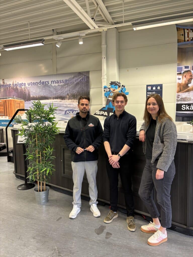 Tre personer står innendørs ved siden av en potteplante og utstillingsdisker, med reklameplakater på veggen bak dem.