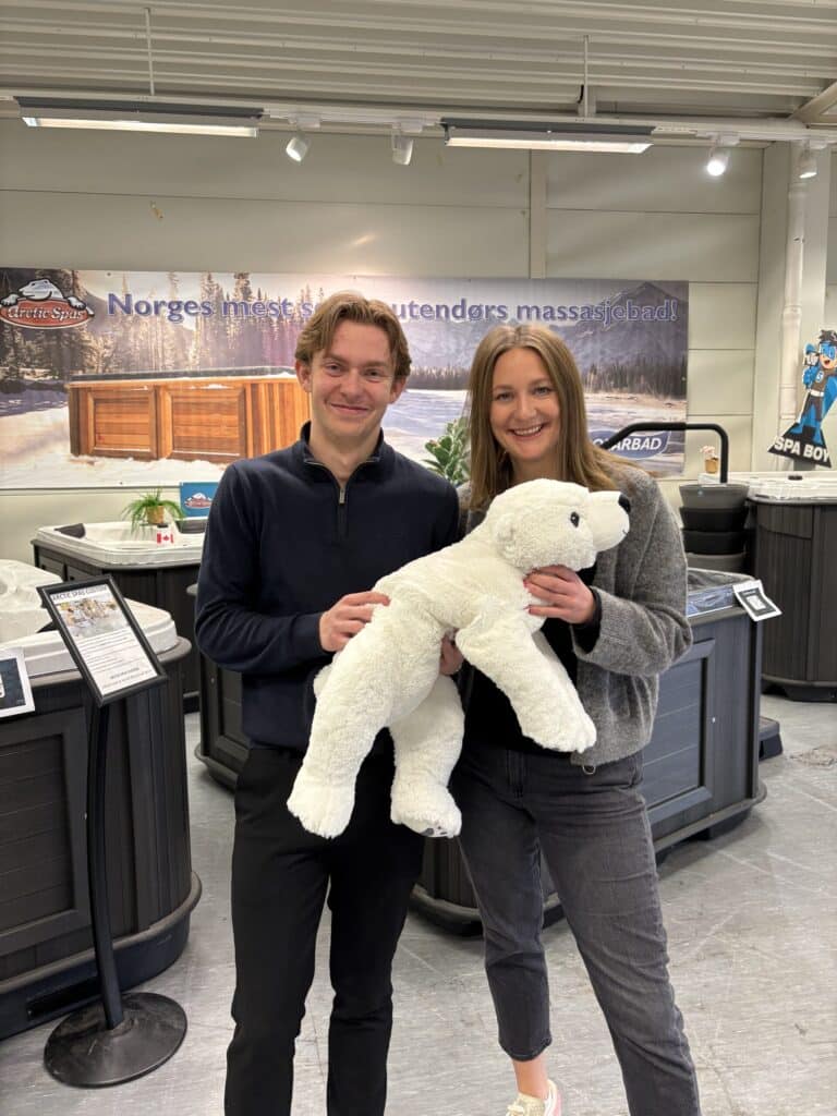 To personer står innendørs og smiler til kameraet, mens den ene holder en hvit isbjørn. I bakgrunnen ser man boblebad og informasjonsskilt.