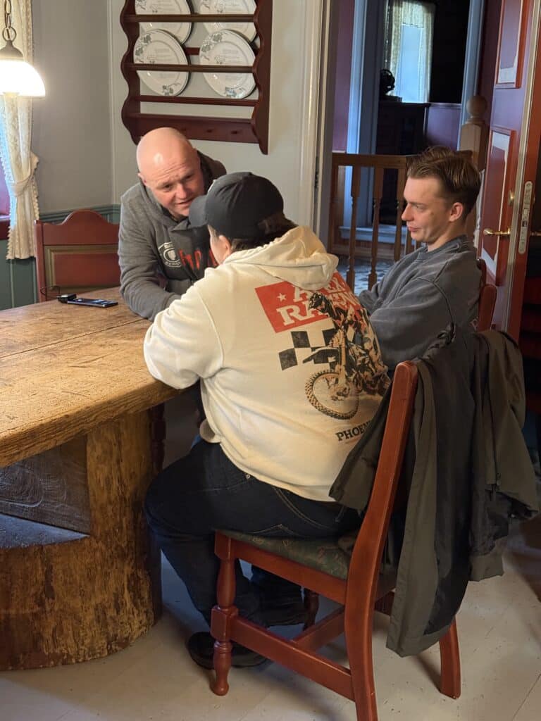Tre personer sitter rundt et trebord innendørs og snakker sammen. En av dem er vendt bort iført hettegenser, mens de to andre sitter vendt mot ham og lytter oppmerksomt.