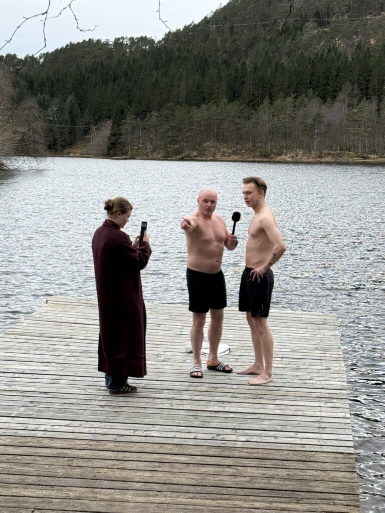 Tre personer står på en trebrygge ved en innsjø; to menn i badetøy ser ut til å være i et intervju, mens en kvinne i morgenkåpe filmer dem med telefonen sin.