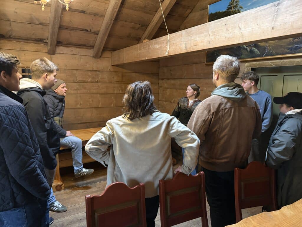 En gruppe mennesker står i et rustikt trerom og lytter til en kvinne som snakker, mens en person sitter på et bord.