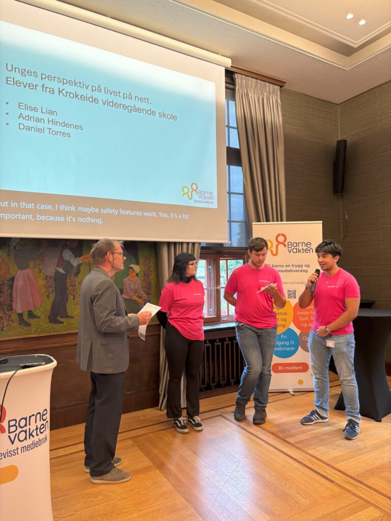 Fire personer står fremst i rommet, tre av dem iført matchende rosa skjorter, og presenterer foran en skjerm med navn og norsk tekst på et arrangement i Barnevakten.