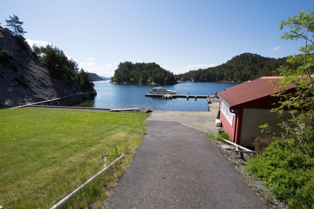 Bilde-241-of-244-scaled En asfaltert sti fører til en brygge med båter på en rolig innsjø, omgitt av gressplener, trær, svaberg og en rød bygning til høyre under en klar himmel.