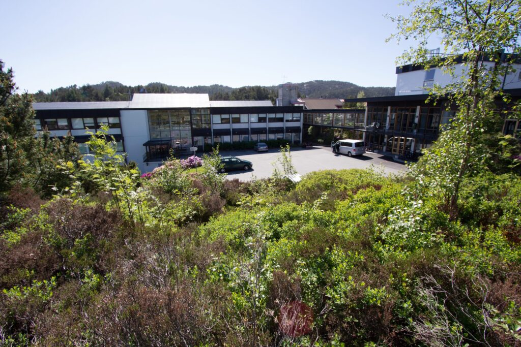 En lav, moderne bygning med store vinduer sees bak en parkeringsplass med to biler, omgitt av trær og grønne busker på en solrik dag.