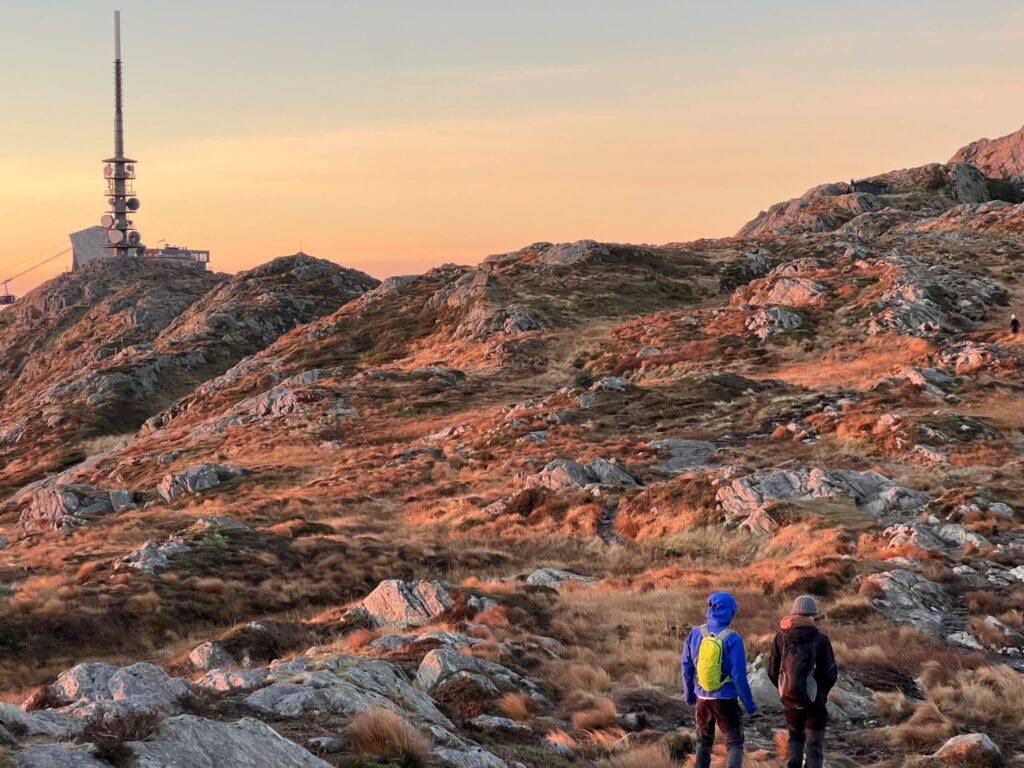 Bilde2-scaled To personer med ryggsekker går på en steinete sti gjennom kupert terreng i solnedgang, med en stor antennestruktur synlig på en ås i det fjerne.