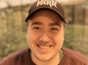 En smilende person med svart Linkin Park-lue og brun skjorte, med en uskarp utendørs bakgrunn.