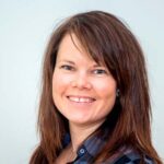 En kvinne med rett, brunt hår og rutete skjorte smiler til kameraet mot en vanlig, lys bakgrunn.