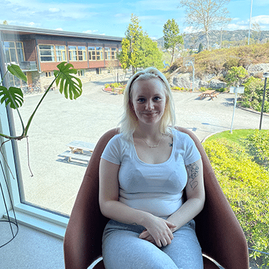 En kvinne med lyst hår sitter i en stol ved et stort vindu, med utsikt over et solfylt uteområde og en bygning i bakgrunnen.