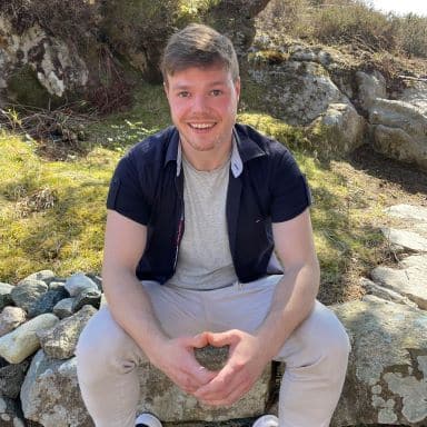 En mann sitter på en steinmur utendørs og smiler til kameraet, med steiner og gress i bakgrunnen i naturlig sollys.