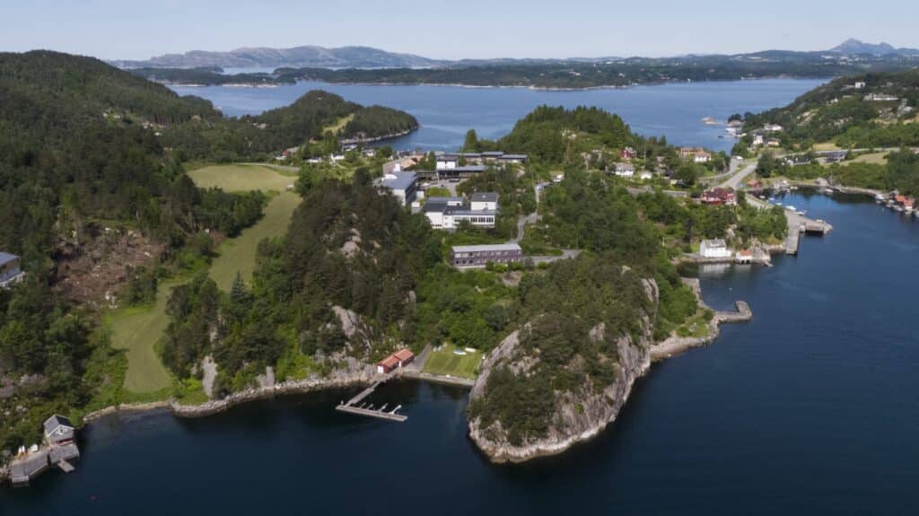 DJI_0009-scaled Flyfoto av en kystlandsby omgitt av grøntområder, med hus, brygger og svaberg langs strandlinjen, og øyer i det fjerne.