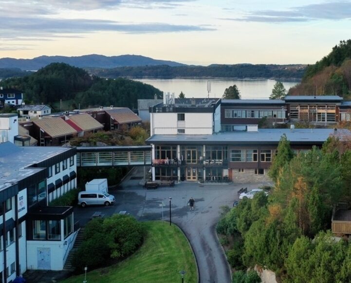 Et skole- eller institusjonskompleks med flere bygninger, omgitt av grøntområder, har utsikt over en rolig innsjø og skogkledde åser under en delvis overskyet himmel.