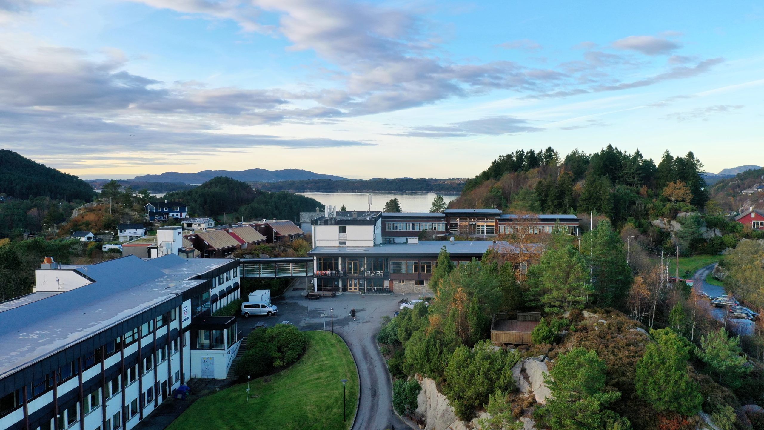 Flyfoto av et skolekompleks omgitt av trær, hus og fjell, med en vannflate synlig i bakgrunnen under en delvis overskyet himmel.