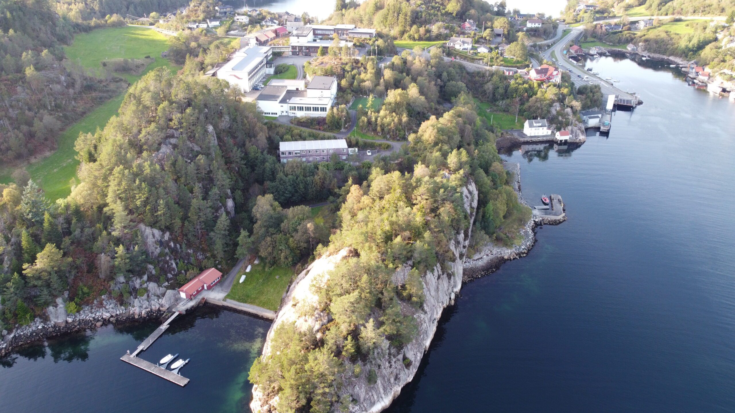 Flyfoto av en skogkledd halvøy med bygninger, brygger og stille vann, omgitt av grønne åser og spredte hus.