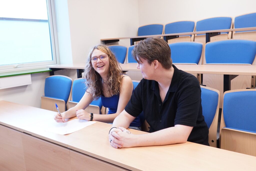 To personer sitter i et klasserom med blå stoler; den ene holder en penn og et papir mens han smiler, den andre ser på dem.