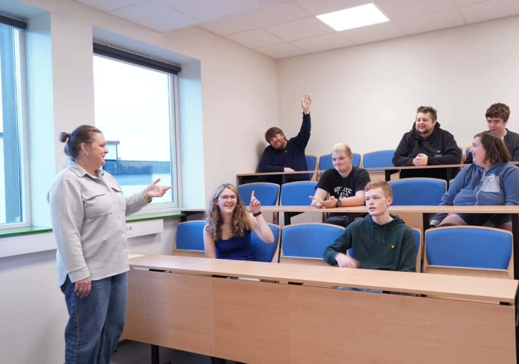 En kvinne står foran i et lite klasserom og gestikulerer mens flere elever sitter på stoler i flere etasjer, og noen av dem rekker opp hånden for å delta.
