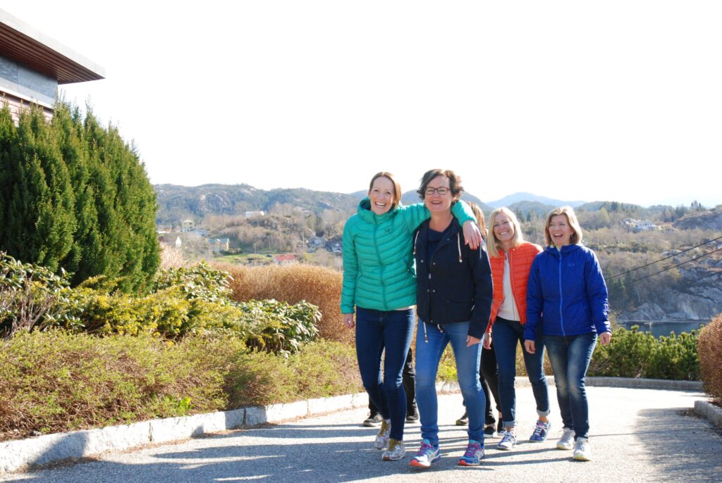 DSC_1049-scaled Fire kvinner i jakker går sammen på en asfaltert sti utendørs, smilende, med busker og åser i bakgrunnen under en klar himmel.
