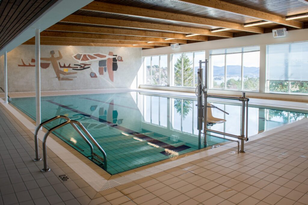 DSC_4330-scaled Innendørs svømmebasseng med heis for bedre tilgjengelighet, flislagt terrasse, store vinduer med utsikt over utendørsområdet og abstrakt veggkunst.