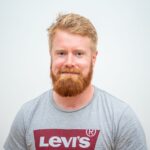 En mann med rødt hår og skjegg, iført en grå Levi's T-skjorte, står mot en vanlig hvit bakgrunn.