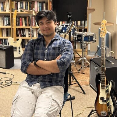 En person sitter med armene i kors i et musikkrom omgitt av gitarer, en forsterker og et trommesett, med bokhyller i bakgrunnen.