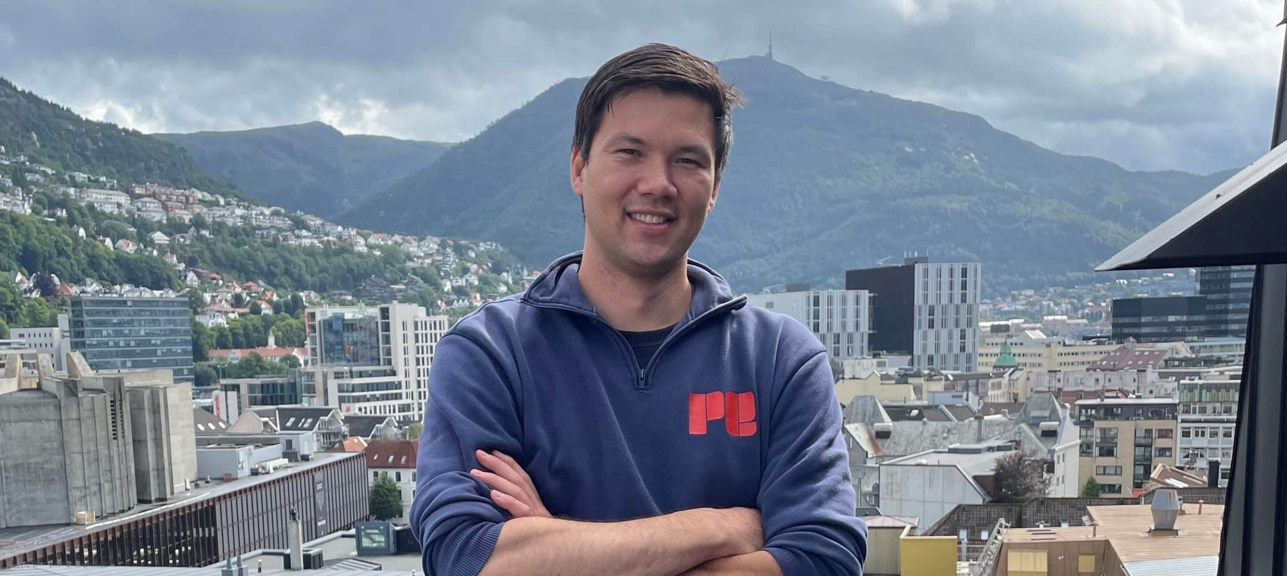 En mann står med armene i kors på et hustak med utsikt over en by med fjell i bakgrunnen på en overskyet dag.