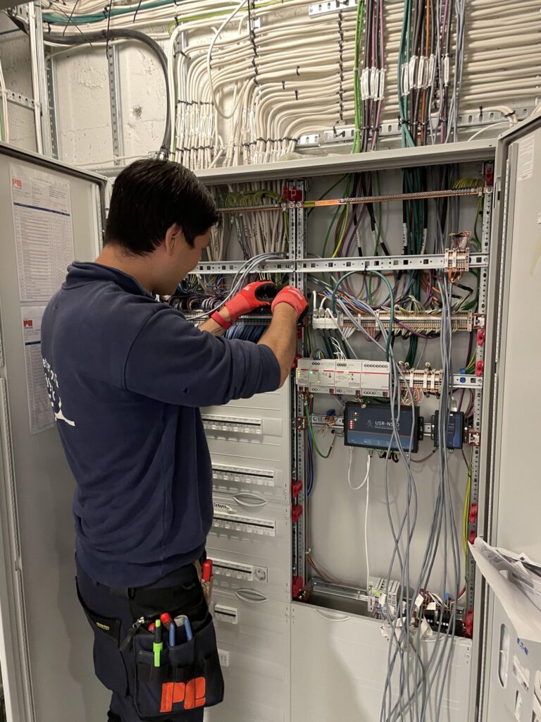 En tekniker med hansker på arbeider med ledninger inne i et åpent elektrisk panel, med verktøy festet til buksene og flere kabler synlige over hodet og inne i skapet.