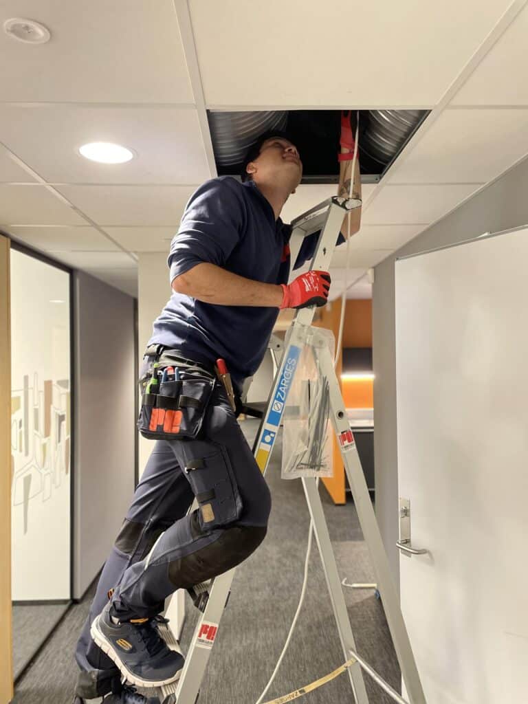 En arbeider på en stige inspiserer eller reparerer en luftkanal i taket på et kontor, iført hansker og med verktøy i beltet.