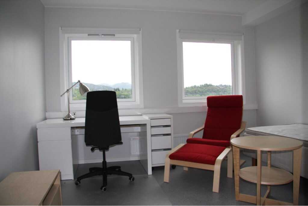 Et minimalistisk kontor med hvitt skrivebord, svart kontorstol, rød lenestol, lite rundt bord, lampe og to store vinduer med utsikt til det fri.