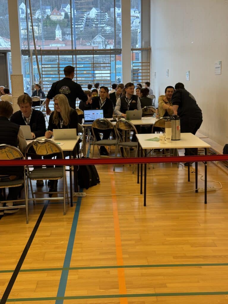 FM UB_2026 (10) I et stort rom med høye vinduer og tregulv sitter flere grupper ved bord med bærbare datamaskiner og deltar i en felles aktivitet eller hendelse.