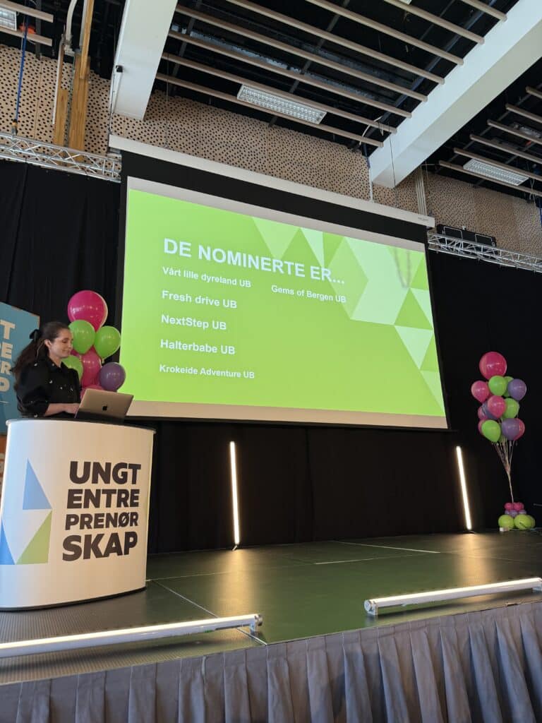 FM UB_2026 (12) En kvinne står på et podium ved siden av en storskjerm som viser en liste over nominerte selskaper på et arrangement, med fargerike ballonger i bakgrunnen.