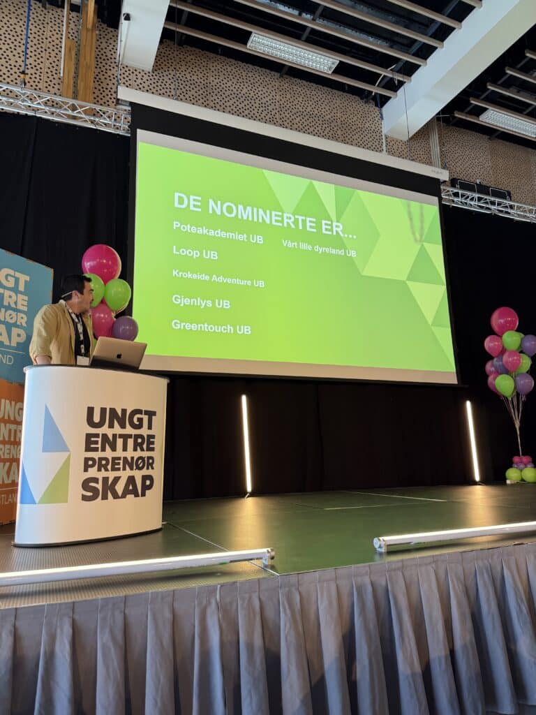 FM UB_2026 (13) En person står på et podium på scenen ved siden av en skjerm som viser en liste over nominerte grupper under overskriften "DE NOMINERTE ER..." på et arrangement med ballonger i bakgrunnen.