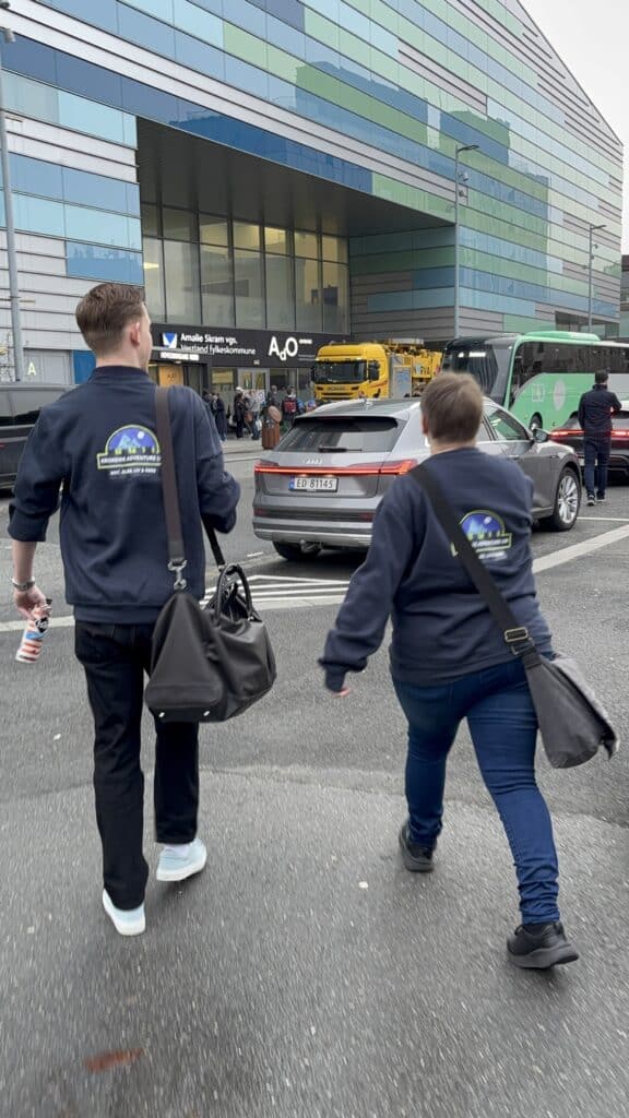 FM UB_2026 (2) To personer med mørke vesker går mot en bygning med skiltet "A03", mens biler og en buss venter på gaten utenfor.