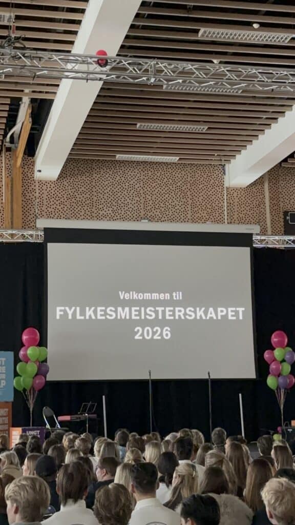 FM UB_2026 (3) Et stort publikum står foran en skjerm som viser "Velkommen til FYLKESMEISTERSKAPET 2026" i et moderne innendørs lokale, med fargerike ballonger på hver side av scenen.