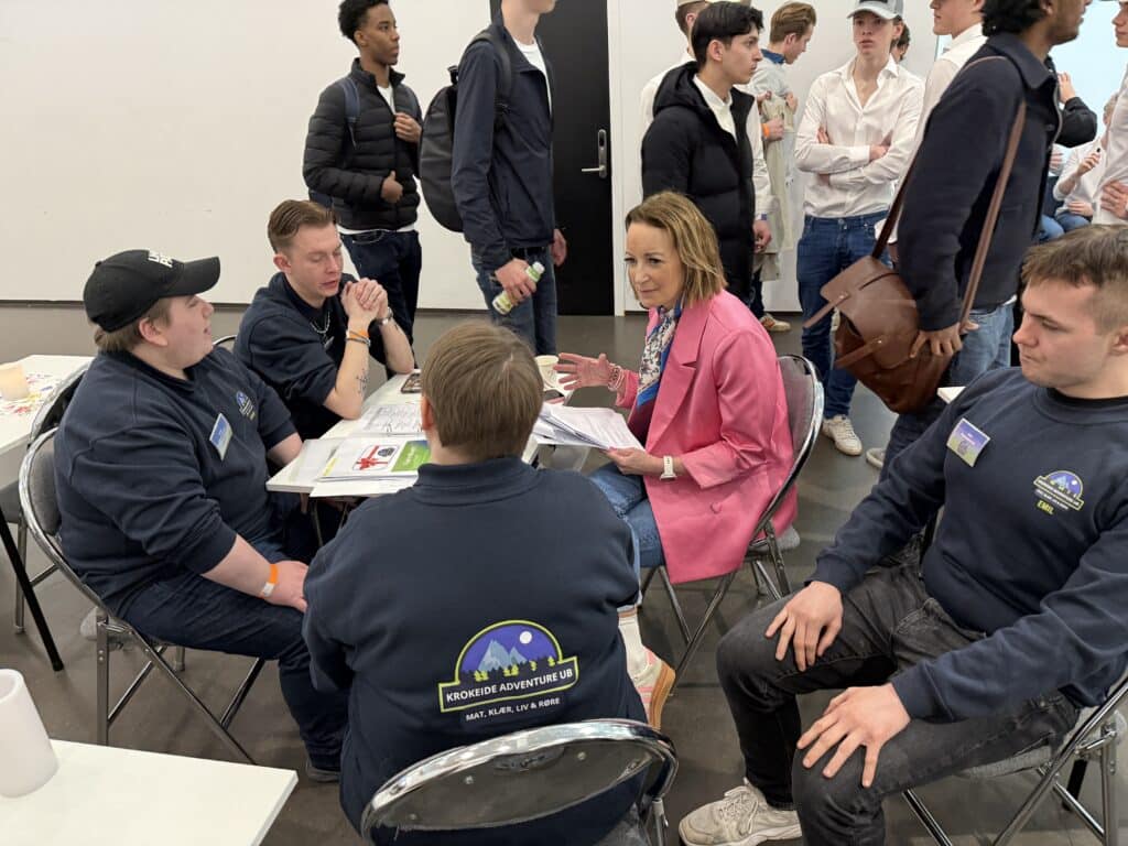 FM UB_2026 (5) En kvinne i rosa blazer snakker med en gruppe unge menn i matchende marineblå skjorter som sitter rundt et bord, mens andre står og observerer i bakgrunnen.