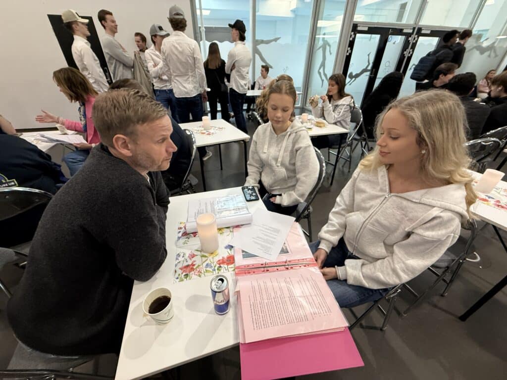 FM UB_2026 (7) Tre personer sitter ved et bord med menyer og drinker i en travel kafé eller restaurant, omgitt av andre gjester og ansatte i hvite uniformer.