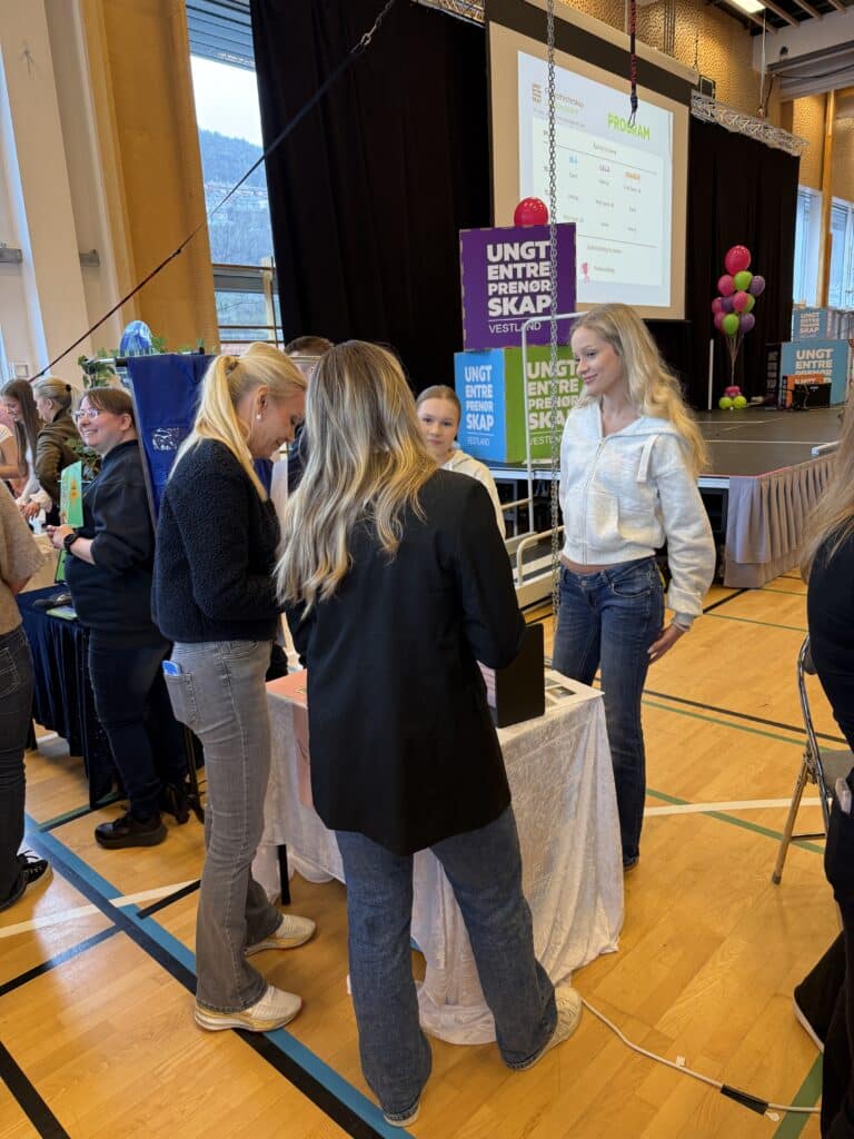 FM UB_2026 (9) Fire unge kvinner står rundt et bord på et innendørsarrangement med bannere, ballonger og en presentasjonsskjerm i bakgrunnen.