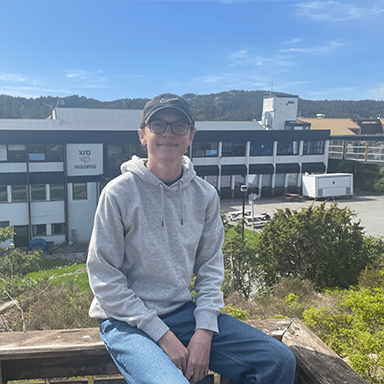 Filip_2024_1405 En person iført grå hettegenser, blå jeans og svart caps sitter på et trerekkverk utendørs med en bygning og trær i bakgrunnen under en blå himmel.