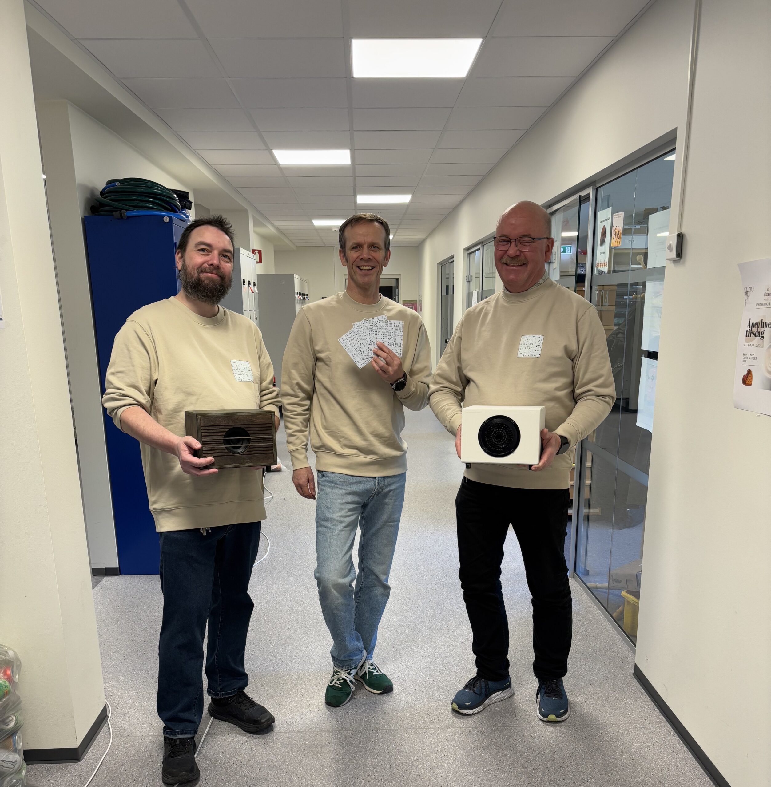 Tre menn i beige gensere står i en gang; den ene holder en trekasse, den andre holder kort, og den tredje holder en hvit eske med en rund åpning.