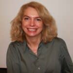 En kvinne med skulderlangt, blondt hår og grå skjorte smiler foran en ensfarget bakgrunn.