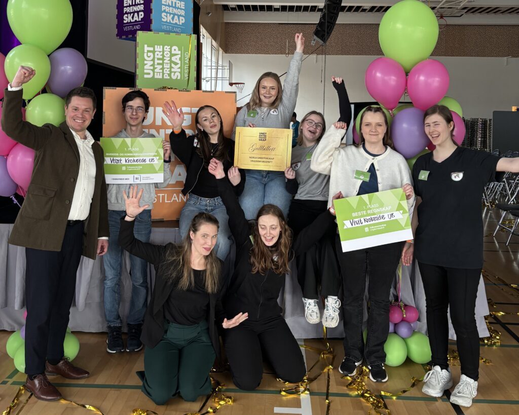 En gruppe på ni personer poserer innendørs med skilt, ballonger og et sertifikat for å feire en prestasjon i et pyntet eventlokale.