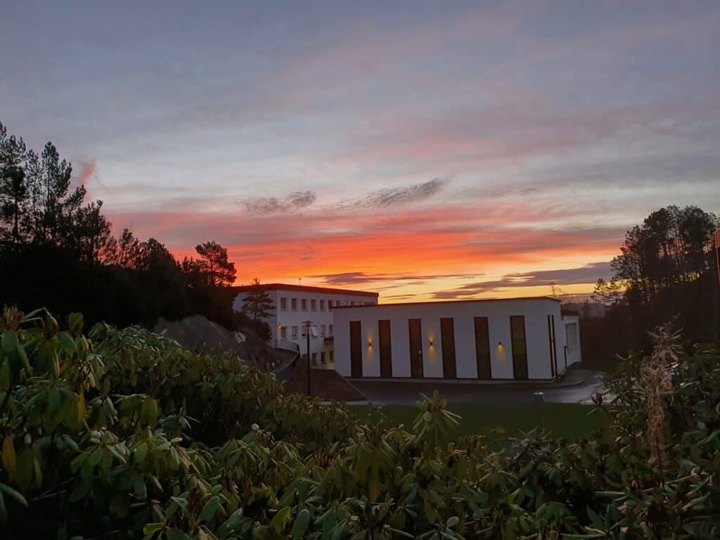 En moderne, hvit bygning ligger mot en dramatisk solnedgangshimmel med oransje og rosa skyer, omgitt av trær og grøntområder.