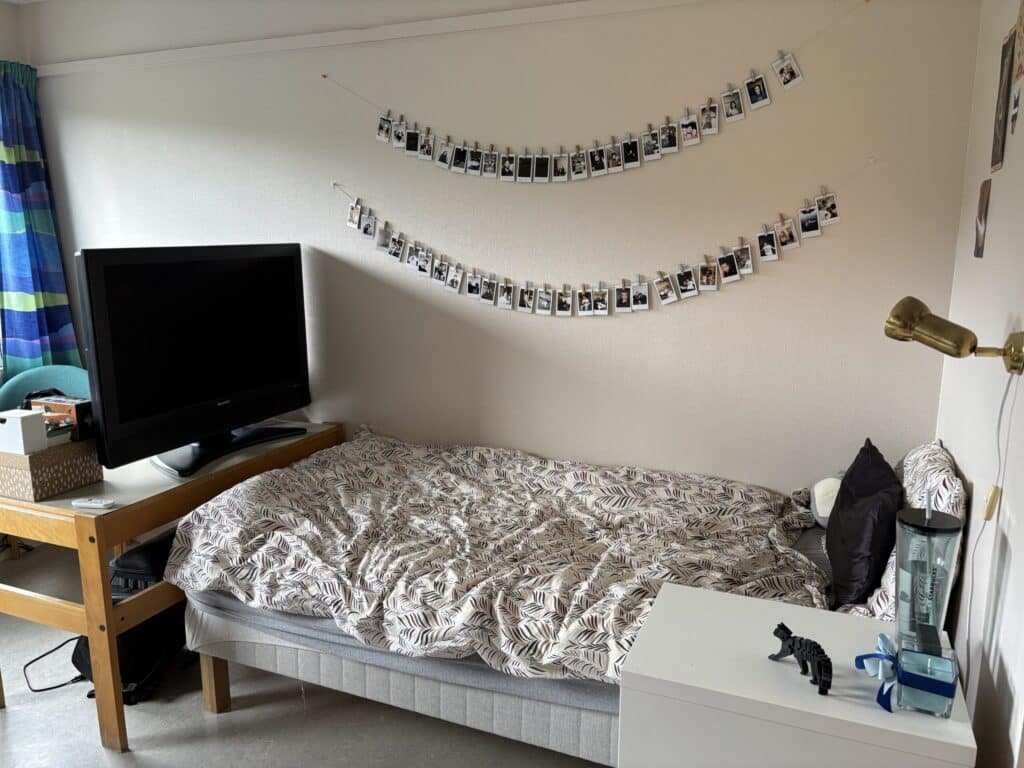 En pent oppredd seng med mønstret sengetøy står mot en vegg dekorert med tre rader med opphengte øyeblikksbilder, ved siden av en TV på et trebord og en hvit kommode med småting.