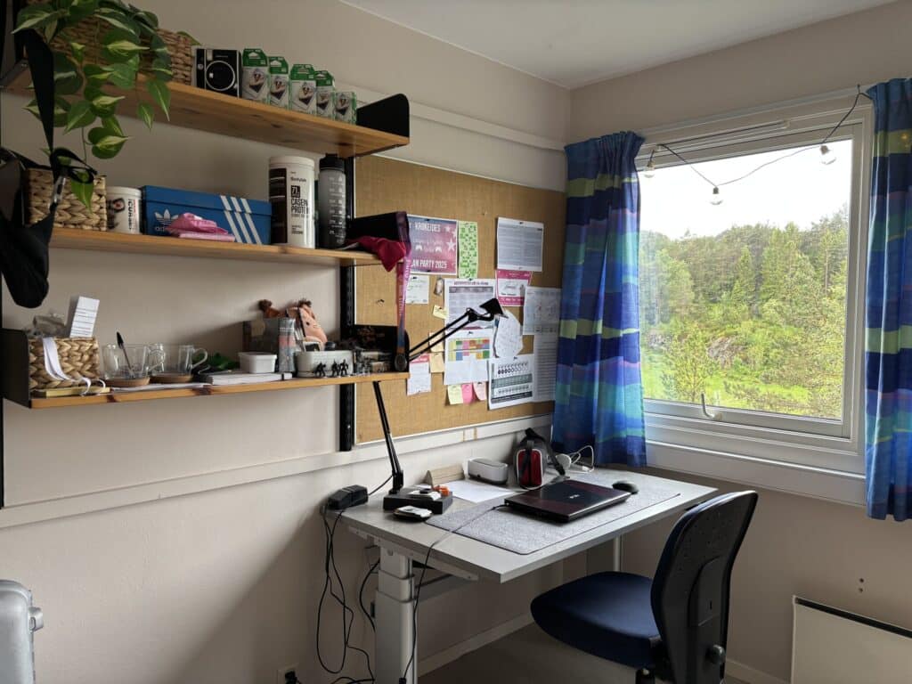 Et hjemmekontor med skrivebord, stol, bærbar PC, lampe, hyller med ulike gjenstander, en korktavle med notater og et vindu med blå gardiner med utsikt over grønne omgivelser.