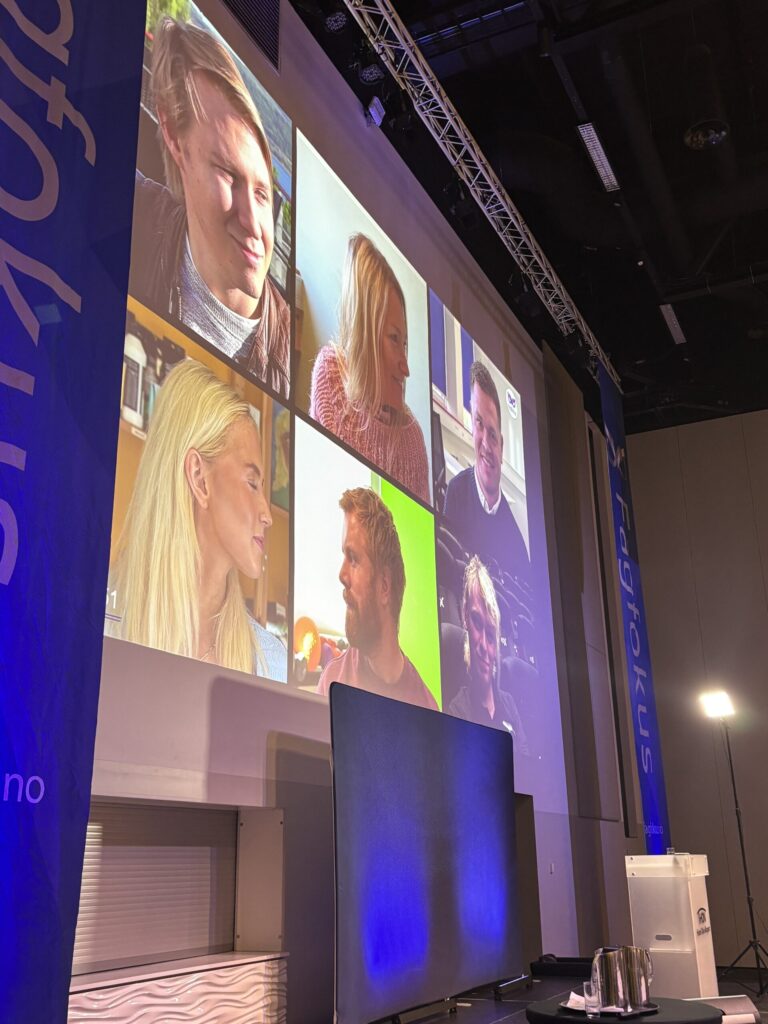 En stor skjerm viser en videosamtale med seks personer i et konferanserom, med blå bannere og scenelys synlig.